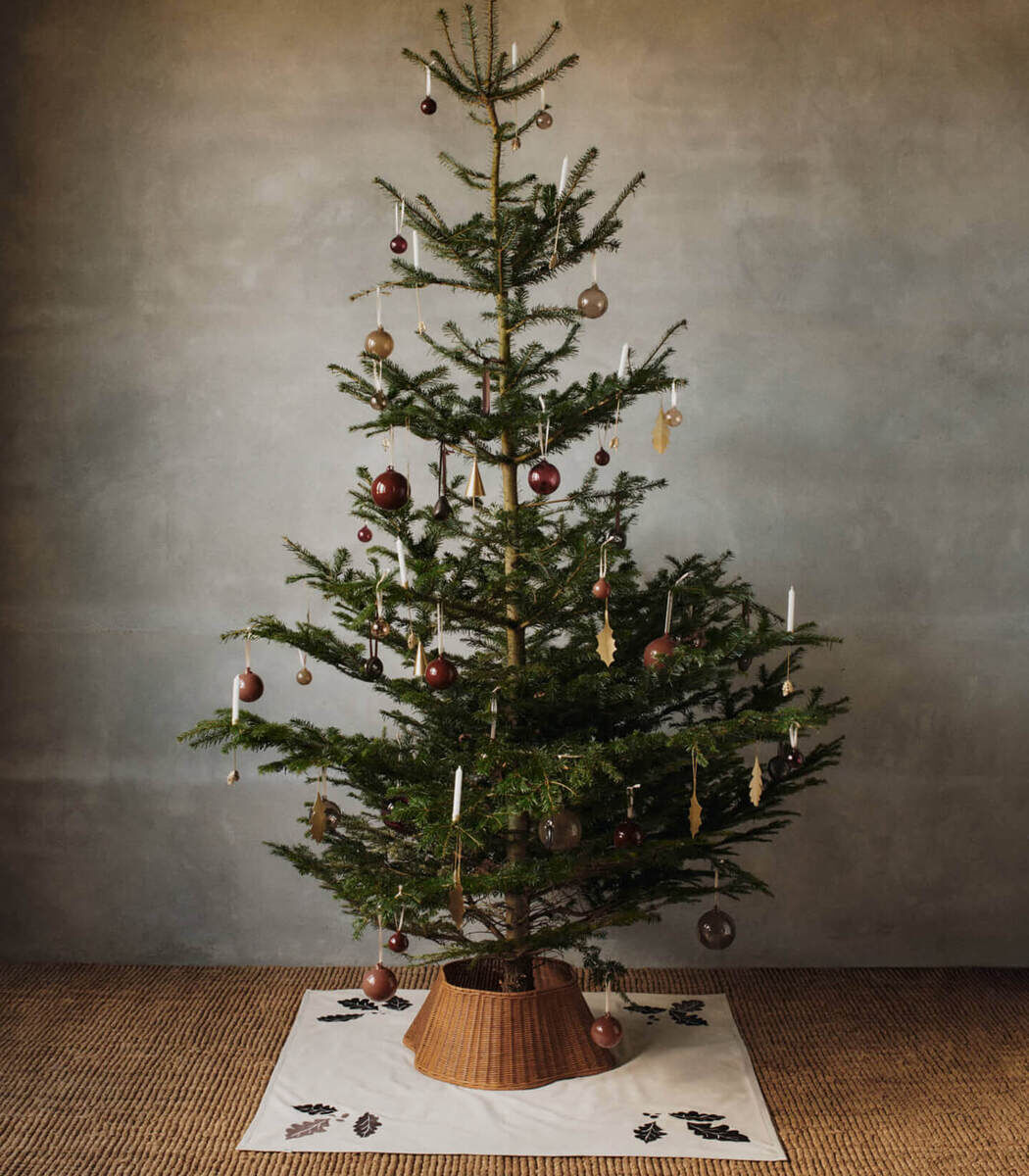Auf dem Bild ist ein natürlich geschmückter Weihnachtsbaum auf einer weißen Christbaumdecke mit Eichenblatt- und Eichelmuster zu sehen. Der Baum steht in einem schlichten Raum mit grauer Wand und Teppichboden. Die Decke betont die festliche Atmosphäre und schützt den Boden.