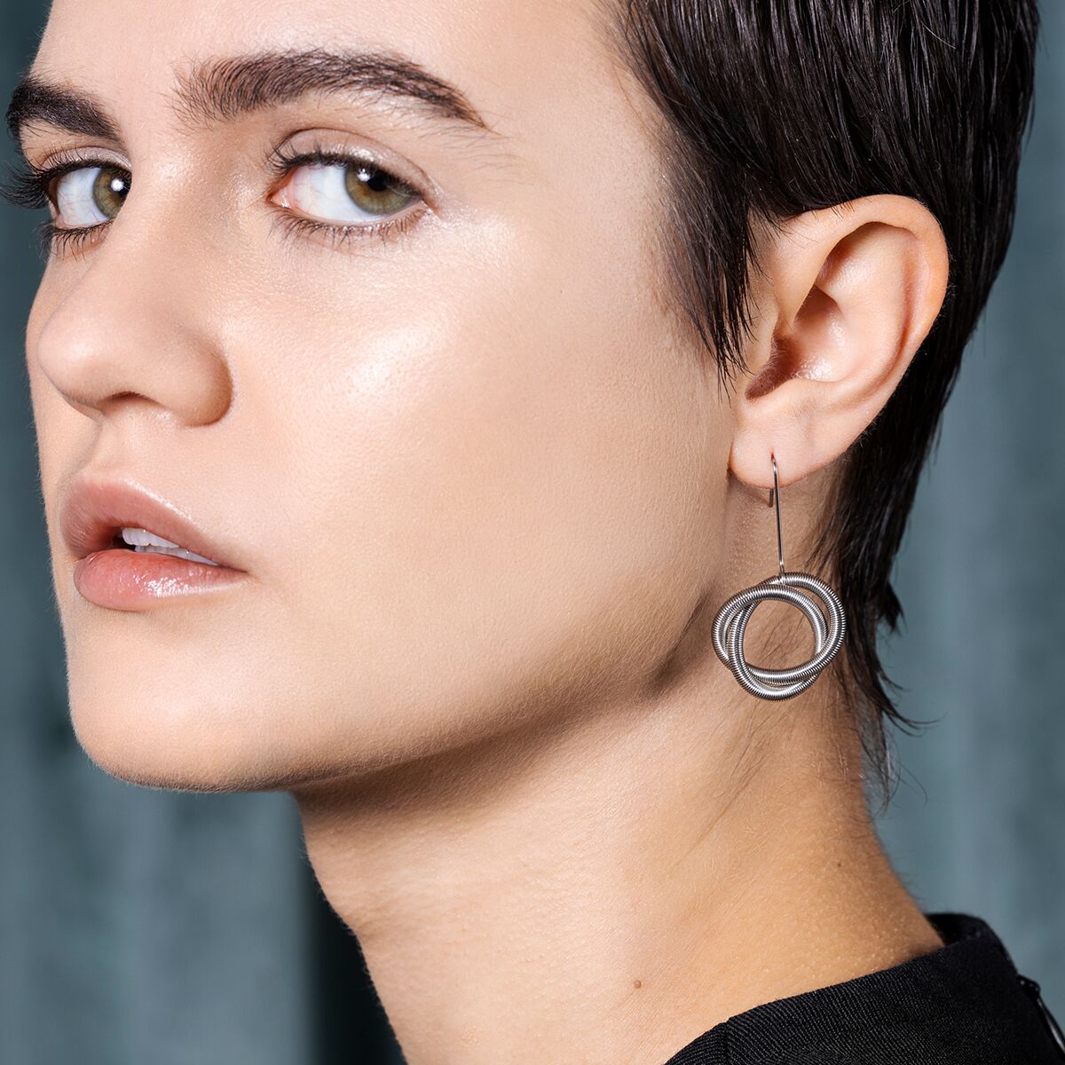 Das Bild zeigt eine junge Person mit kurzen Haaren, die einen auffälligen, silberfarbenen Ohrring mit rundem, geometrischen Design trägt. Der Hintergrund ist unscharf in blauen und grauen Farbtönen gehalten. Der Fokus liegt klar auf dem modernen Schmuckstück am Ohr.