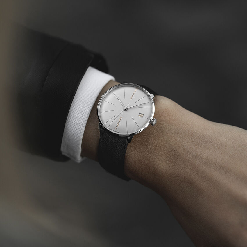 Nahaufnahme einer eleganten Armbanduhr ZEIT-Sonderedition »Meister fein Kleine Automatic« von Junghans am Handgelenk einer Person. Die Uhr hat ein weißes Zifferblatt mit minimalistischen Markierungen, ein silbernes Gehäuse und ein schwarzes Armband.