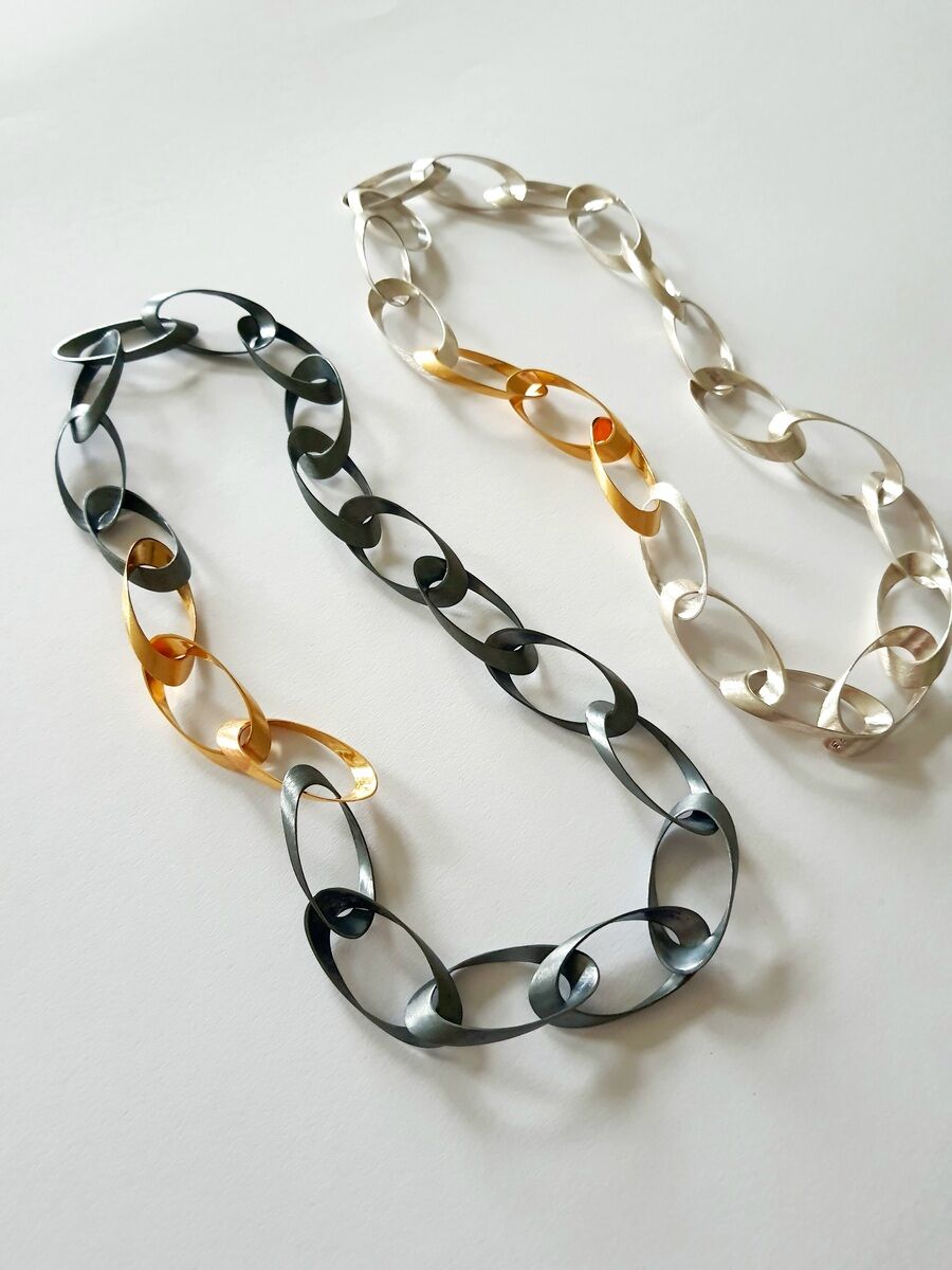 Zwei Halsketten mit Kettengliedern, eine mit Silber- und Goldakzenten, die andere in Silber, auf weißem Hintergrund.