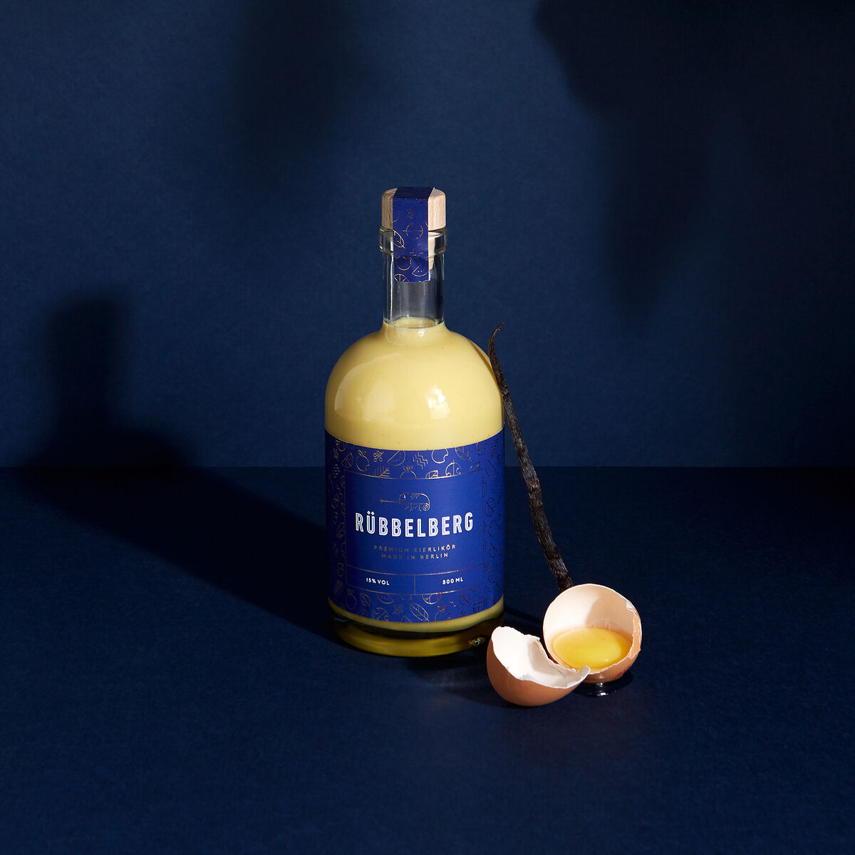Eine Flasche Premium Eierlikör von Rübbelberg steht vor dunkelblauem Hintergrund, begleitet von einer geöffneten Vanilleschote und halben Eierschalen.
