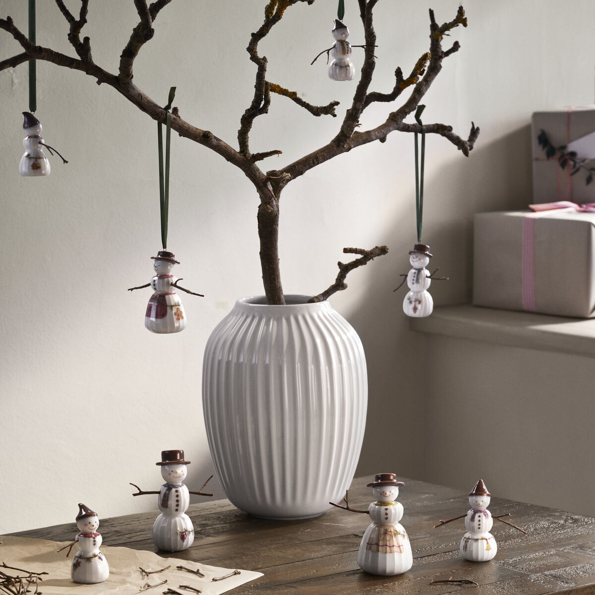 Mehrere Schneefrau-Keramikfiguren der Serie »Hammershøi« stehen auf einem Holztisch und hängen an Ästen in einer weißen Vase. Die Szene ist liebevoll mit Winterdekoration arrangiert und vermittelt eine gemütliche, skandinavische Weihnachtsstimmung.