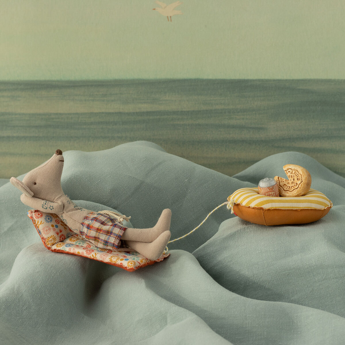 Zwei Stoffmäuse, eine auf einer Luftmatratze, die andere in einem kleinen Boot, posieren vor einem maritimen Hintergrund mit Wellenmustern.