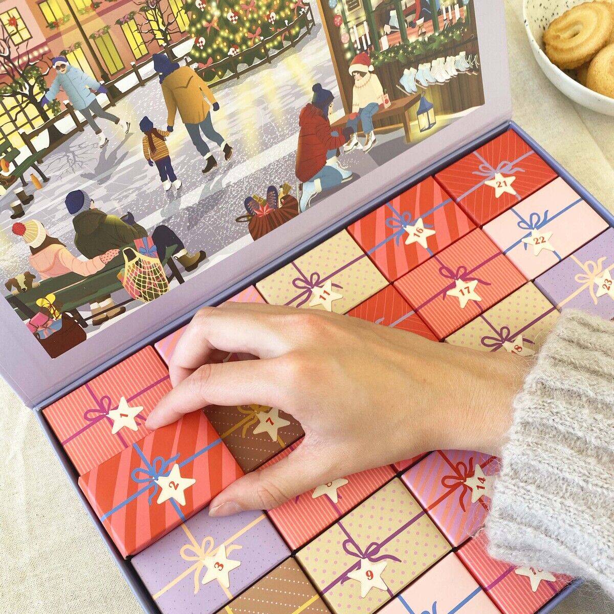 Ein bunter Puzzle-Adventskalender »Skate & Coffee« von VISSEVASSE liegt geöffnet auf einem Tisch. Die Box zeigt eine winterliche, belebte Straßenszene im Deckel und 25 kleine, als Geschenke gestaltete Schachteln. Eine Hand öffnet gerade eine der Schachteln.