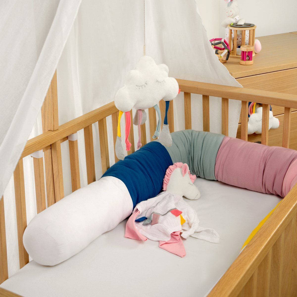 Eine niedliche Spieluhr in Wolkenform hängt über einem Kinderbett, das mit bunten Bettwurstkissen ausgestattet ist. Die Wolkenspieluhr hat bunte Bänder, die herabhängen, und der Raum ist in hellen Farben dekoriert.