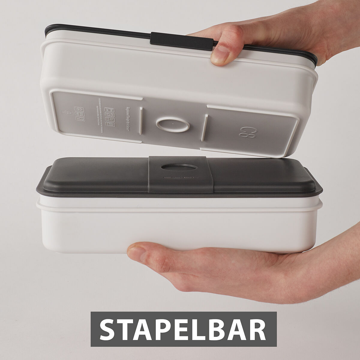 Eine doppelstöckige, stapelbare Lunchbox aus weißem und schwarzem Plastik wird von zwei Händen gehalten. Das untere Fach ist weiß, während das obere Fach schwarz ist. Auf der Verpackung steht »STAPELBAR«.