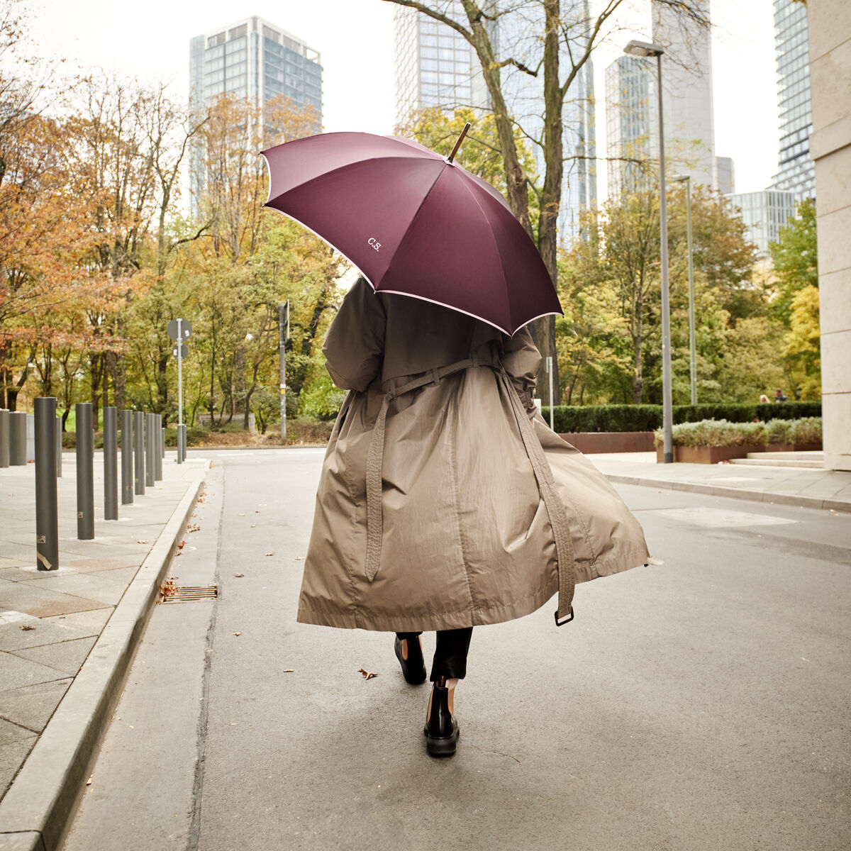 Eine Person mit beigefarbenem Trenchcoat hält einen bordeauxroten Regenschirm »Norfolk Oxford« Berry und geht auf einer mit Laub gesäumten Straße in einer modernen Stadt mit hohen Gebäuden. Der Regenschirm schützt vor leichtem Regen, während herbstliche Bäume den Hintergrund bestimmen.