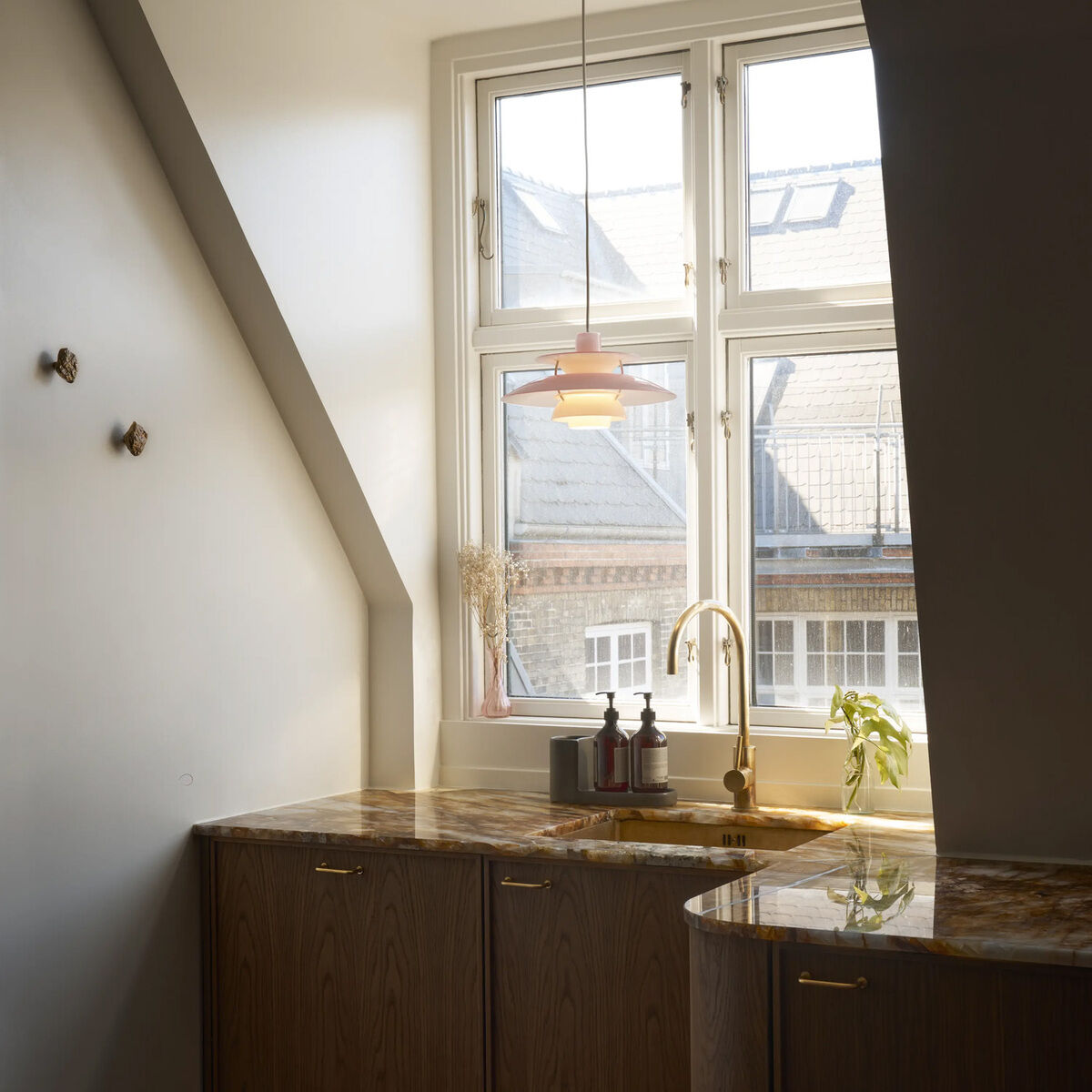 Hängeleuchte »PH 5 320« von Louis Poulsen in Weiß, Pale Rose und Beige hängt vor einem Fenster mit Blick auf Nachbargebäude über einer eleganten Küche mit gemaserten Holzfronten und goldener Spüle, natürlichem Lichteinfall und dekorativen Details.