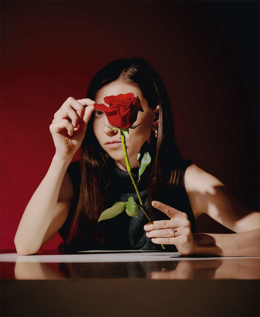 Frau mit langer dunkler Haaren hält eine rote Rose vor ihrem Gesicht, vor einem roten Hintergrund. Die Szene ist geheimnisvoll und schattig beleuchtet.