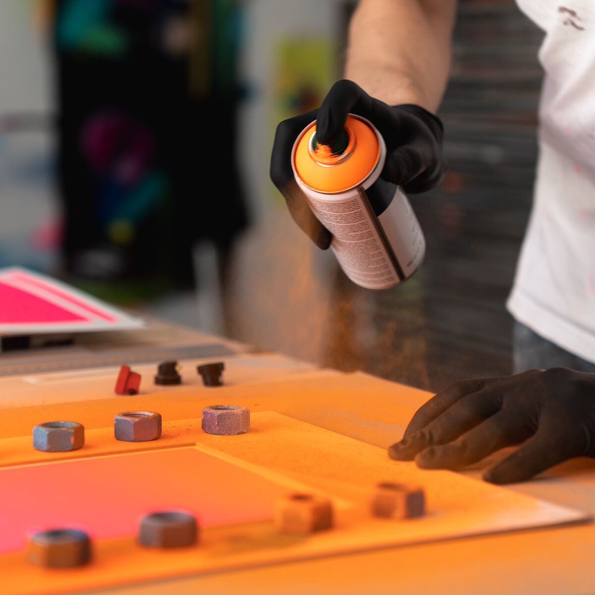 Ein Handschuh tragender Mensch sprüht mit einer Sprühdose orange Farbe auf ein modernes Kunstwerk mit intensiven Pink- und Orangetönen. Im Vordergrund liegen Farbkappen und verschiedene Werkzeuge auf einem Tisch in einem hellen, kreativen Atelier.