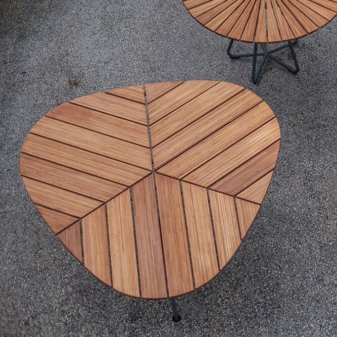 Eine Abbildung von drei Gartentischen der Serie »LEAF«. Die Tische bestehen aus holzartigen Oberflächen mit grauen Mitten und sind rund oder dreieckig geformt, auf einem grauen Kiesboden platziert.