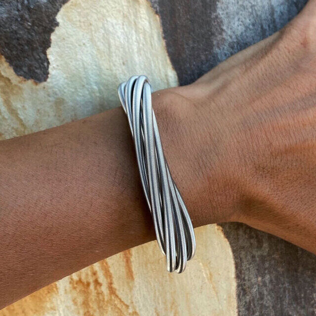 Nahaufnahme eines Handgelenks, das ein elegantes, silbernes Twist-Armband trägt. Der Hintergrund zeigt eine holzartige Textur in Beige-, Weiß- und Brauntönen.