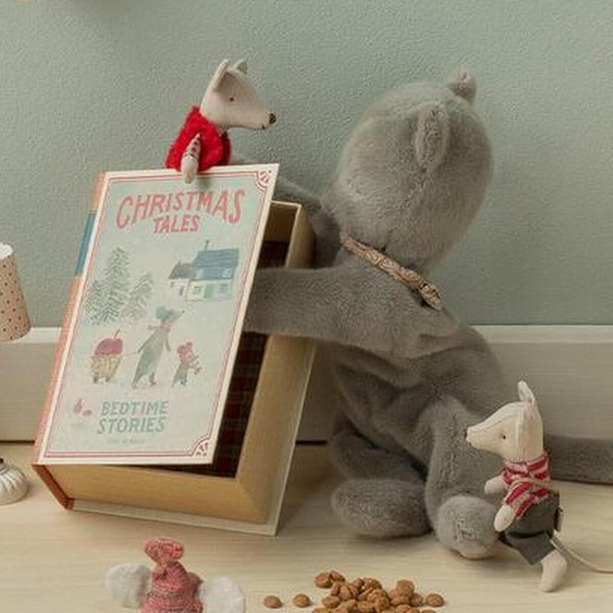 Mehrere Maileg-Stofftiere, darunter ein großes Schwein und verschiedene weihnachtlich gekleidete Mäuse, sitzen auf hellem Boden vor einer mintgrünen Wand. Sie sind um Miniaturmöbel, ein Buch und kleine Dekorationen arrangiert und bilden eine festliche, winterliche Spielszene.