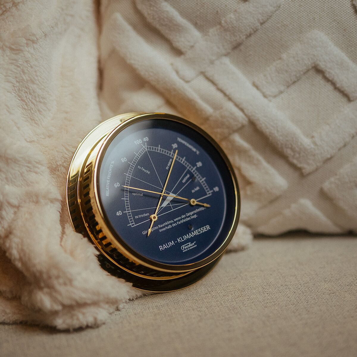 Ein elegantes Raumklimamessgerät der ZEIT Sonderedition aus poliertem Messing mit blauem Ziffernblatt liegt auf einem beigen Kissen mit weißer Struktur im Hintergrund. Das Messgerät zeigt verschiedene Klimaanzeigen wie Temperatur und Luftfeuchtigkeit und dient als stilvolles Wohnaccessoire.