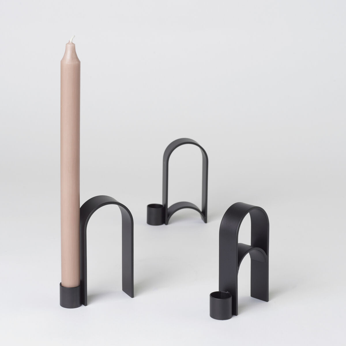 Ein modernes Design-Kerzenhalter-Set aus Metall namens 'Arch' Vol.10 in Schwarz. Der Kerzenhalter verfügt über einen eleganten und minimalistischen Bogenstil. Eine beige Kerze ist in einem der Kerzenhalter platziert. Geeignet für eine Vielzahl von Anlässen, insbesondere als stilvolle Inneneinrichtung.