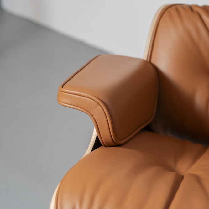 Detailaufnahme eines Lounge Chairs und Ottomans. Der Stuhl ist mit braunem Leder bezogen und hat eine bequeme Polsterung sowie Armlehnen. Der Rahmen besteht aus Holz in einem hellen Farbton, und der Boden ist neutral grau.