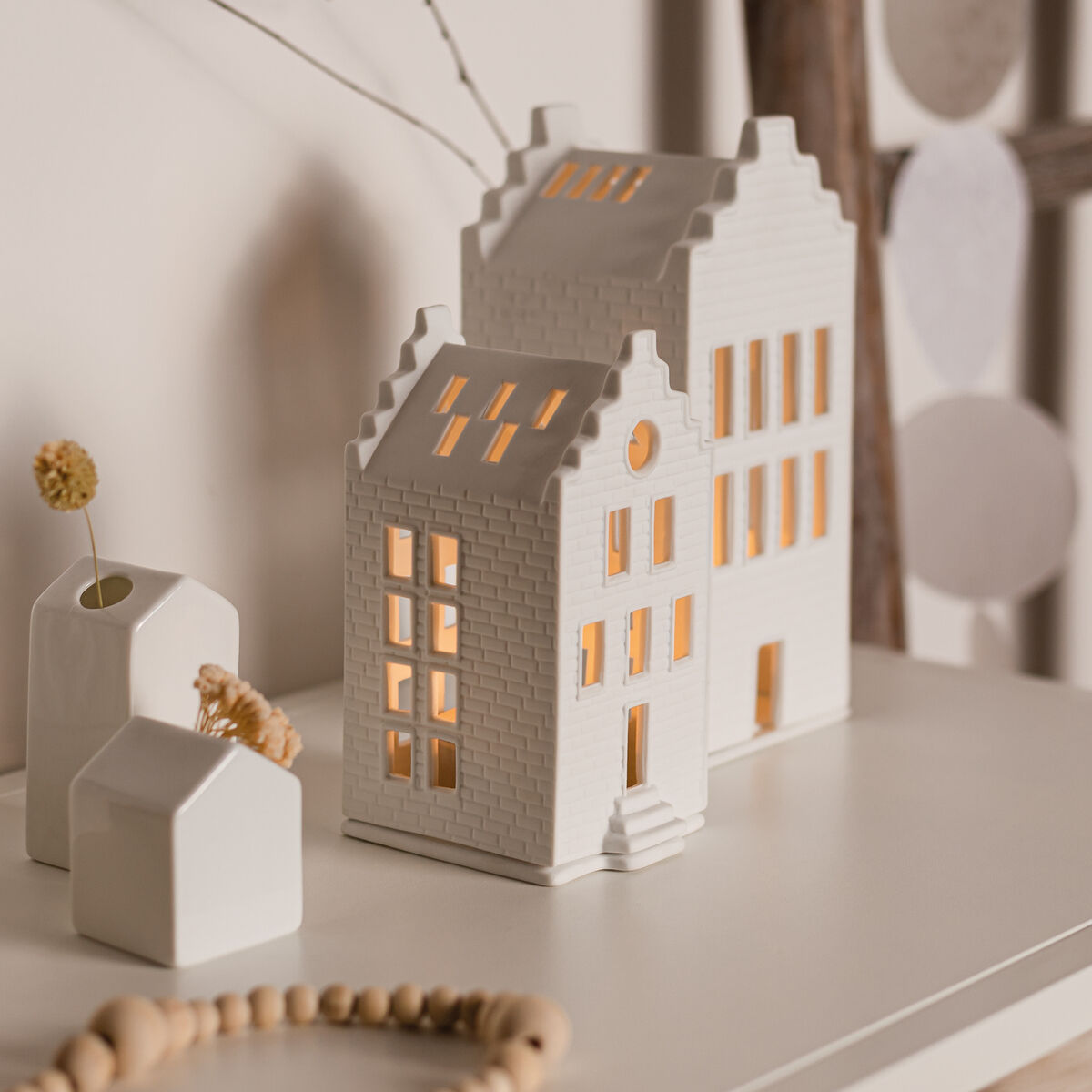 Ein dekoratives, weißes Lichthaus in Form eines kleinen Backsteinhauses steht auf einem hellen Regal. Es ist von zarten, natürlichen Accessoires wie kleinen Vasen, getrockneten Blüten und einer Holzperlenkette umgeben. Das Keramikhaus leuchtet warm durch die Fenster, wodurch eine gemütliche Atmosphäre entsteht.
