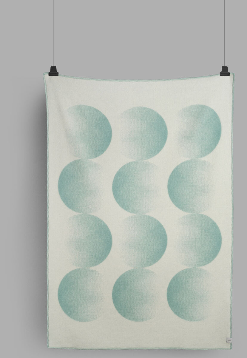 Eine Lammwolldecke mit dem Namen »MOON«, die an einer grauen Wand hängt. Die Decke ist weiß mit mintgrünen Kreismustern.