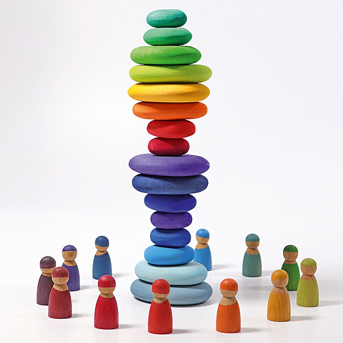 Mehrere flache, farbig lasierte Holzsteine sind in einem hohen, ausbalancierten Turm von Hellgrün über Gelb, Orange, Violett bis zu Blau übereinandergestapelt. Rundherum stehen kleine bunte Holzfiguren in leuchtenden Farben auf weißem Untergrund.