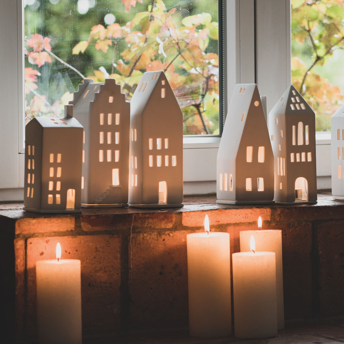 Eine Sammlung von weißen Lichthäusern verschiedener Formen und Größen, die auf einer Fensterbank mit Hintergrundbeleuchtung durch Kerzen stehen. Im Hintergrund sichtbares Fenster mit wenigen grünen und gelben Blättern an einem Baum.