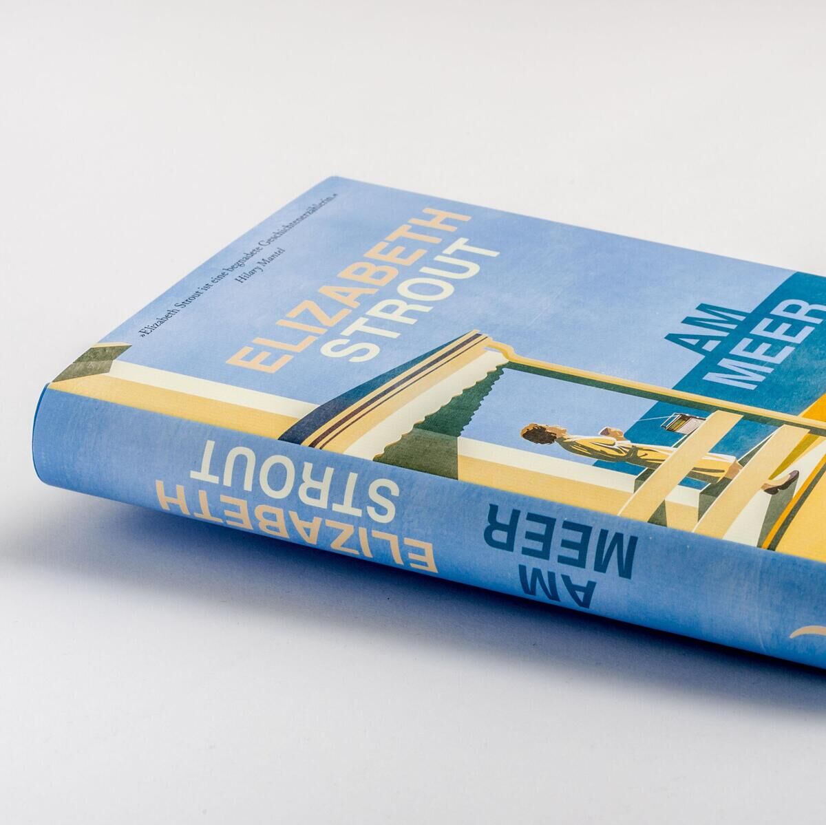 Das Bild zeigt das Buch 'Am Meer' von Elizabeth Strout. Das Buch hat einen blauen und gelben Einband mit einem minimalistischen Strandmotiv. Der Einband zeigt eine sitzende Person, die auf das Meer blickt.