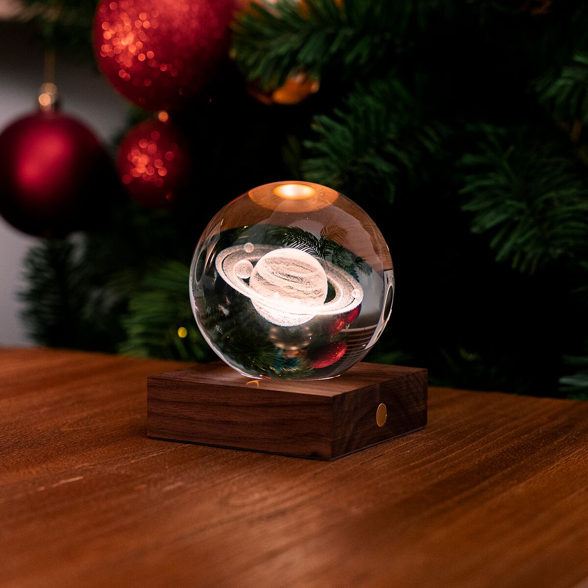Glaskugel mit Saturn-Motiv auf einem Holzsockel, beleuchtet von warmem Licht, vor einem festlich geschmückten Weihnachtsbaum mit roten Kugeln auf einem Holztisch, geeignet als stimmungsvolles Wohnaccessoire oder Geschenk.