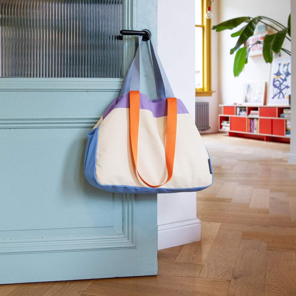 Eine stilvolle Canvas Tasche mit lila, orange und blauen Details hängt an einer tür in einem Raum mit Holzfußboden und dekorativen Elementen.