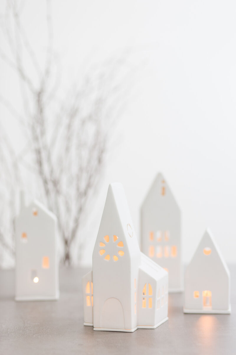 Keramiklichthaus in Form einer kleinen Kirche mit leuchtenden Fensteröffnungen. Zeigt eine minimalistische weiße Keramik mit weichem gelbem Licht, umgeben von weiteren ähnlichen Keramikhäusern und einem unscharfen Hintergrund von kahlen Zweigen. Perfekt als Dekoration für Weihnachten und andere festliche Anlässe.