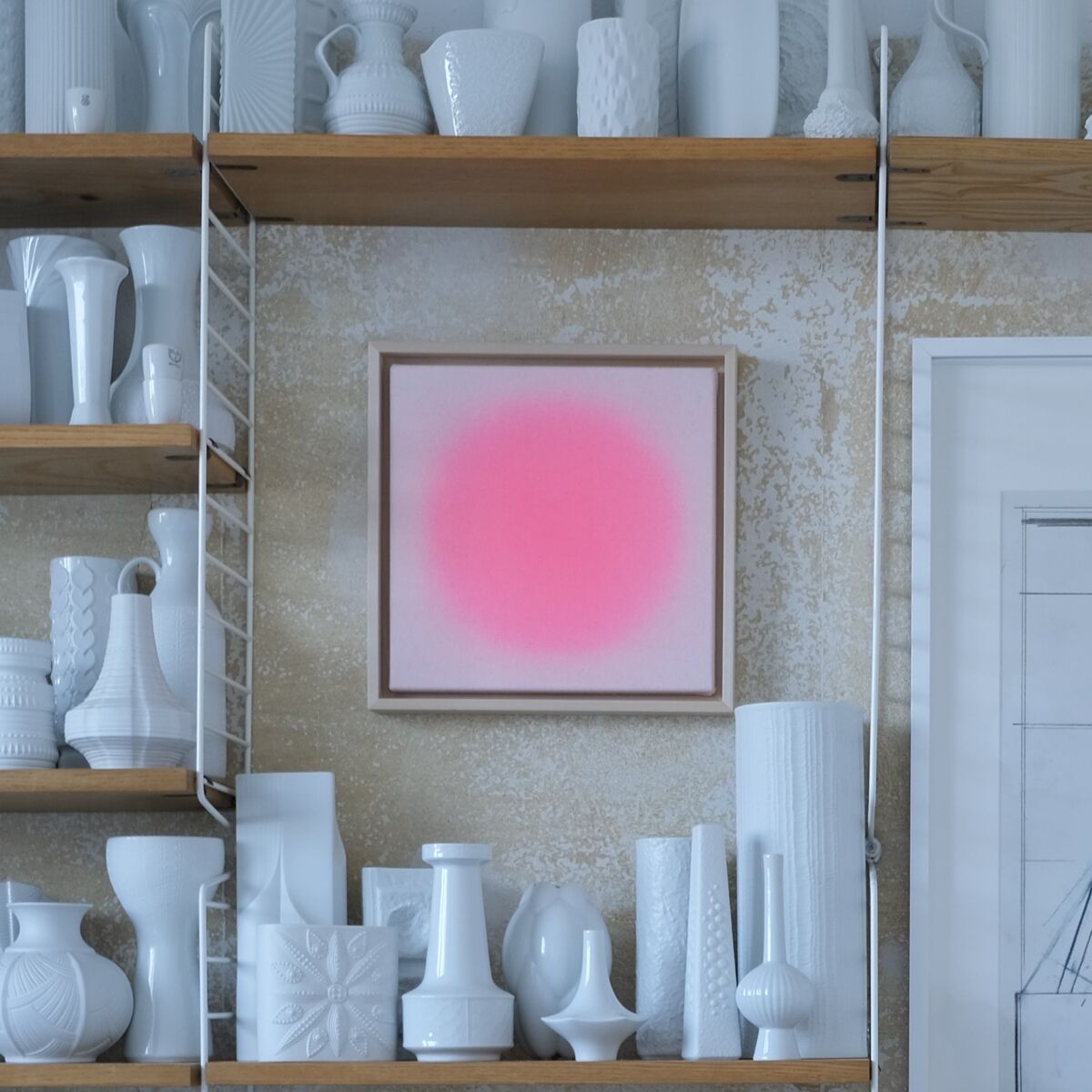 Das Bild zeigt eine stilvolle Wand mit offenem Regal, auf dem zahlreiche weiße Keramikobjekte arrangiert sind. In der Mitte hängt ein leuchtendes Kunstwerk in Pink, die ZEIT-Sonderedition »Sorbet« von Anastasia Benko. Daneben ist ein schwarz-weißes Bild zu sehen. Der Boden ist mit weichem, flauschigem weißem Teppich bedeckt.