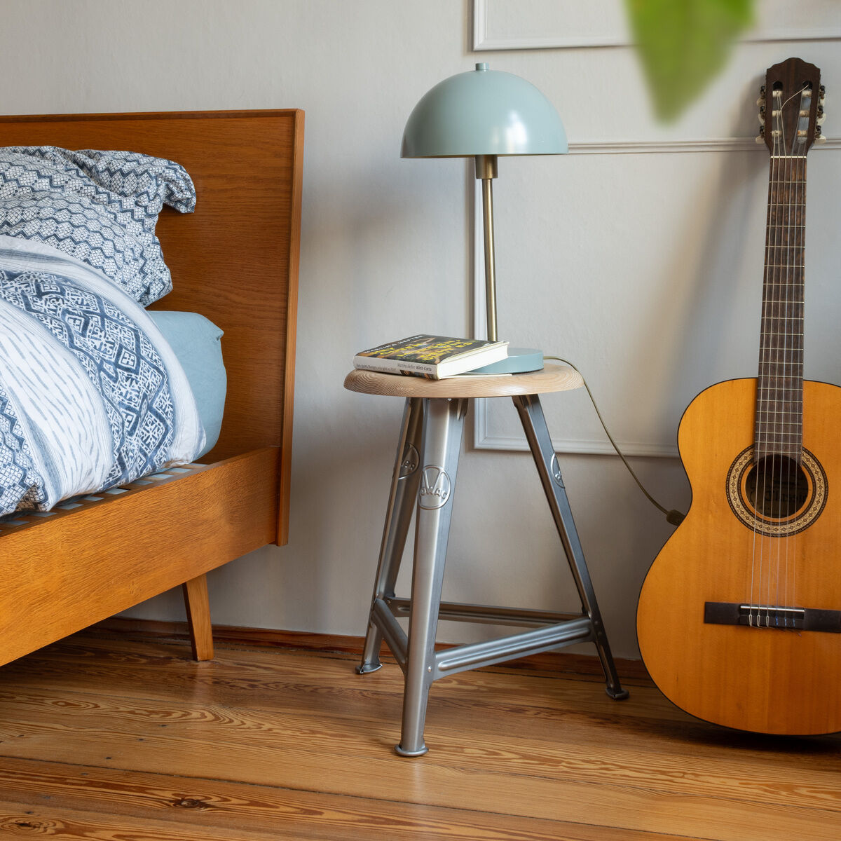 Ein designorientierter Metallhocker mit naturbelassener Esche-Sitzfläche steht als Nachttisch neben einem modernen Holzbett in einem stilvoll eingerichteten Schlafzimmer. Auf dem Hocker liegen ein Buch und eine hellblaue Tischlampe, daneben steht eine klassische Gitarre auf einem Holzboden. Die Szene vermittelt ein gemütliches und modernes Wohnambiente.