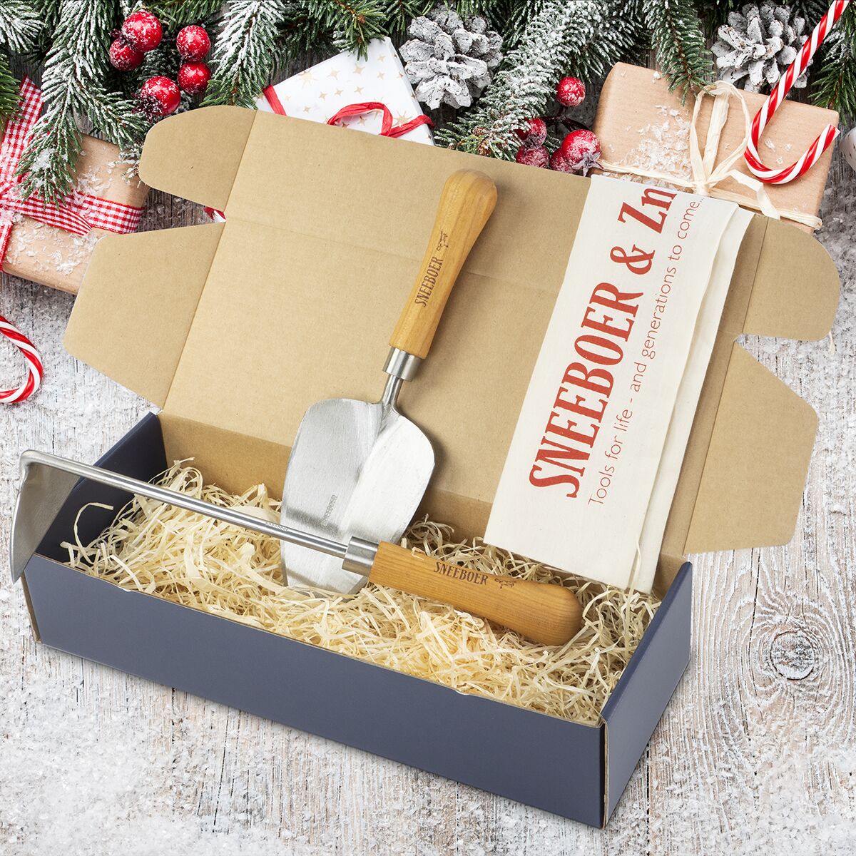 Offene Geschenkbox mit zwei hochwertigen Gartenwerkzeugen von Sneeboer & Zn – einer Pflanzkelle und einem Jäter aus silbrigem Metall mit edlen Kirschholzgriffen – liegt dekorativ auf Stroh. Im Hintergrund Winterdeko mit Geschenkpaketen, Schnee und Tannenzweigen.