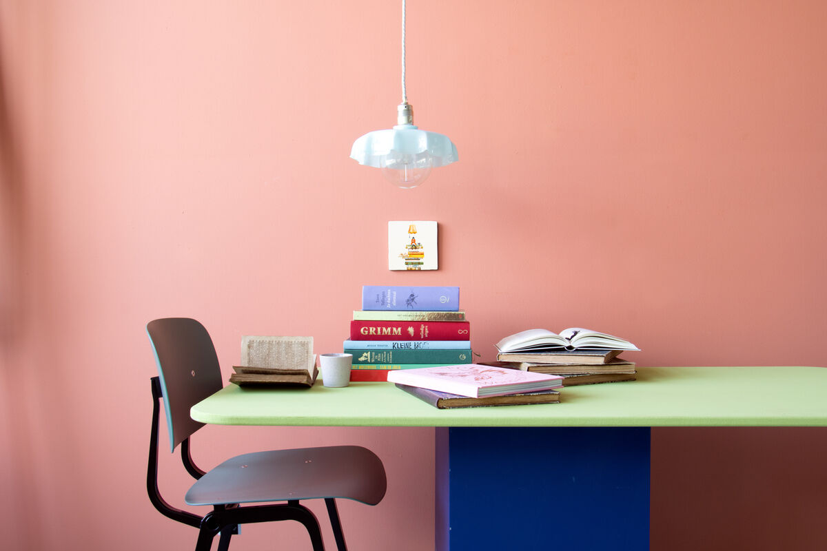 Eine Keramikfliese mit der Aufschrift »Bücherwurm« hängt an einer rosa Wand. Darunter befinden sich ein Schreibtischstuhl, ein stapel Bücher, und ein hängendes Licht. Ein Buch ist geöffnet auf dem Tisch.