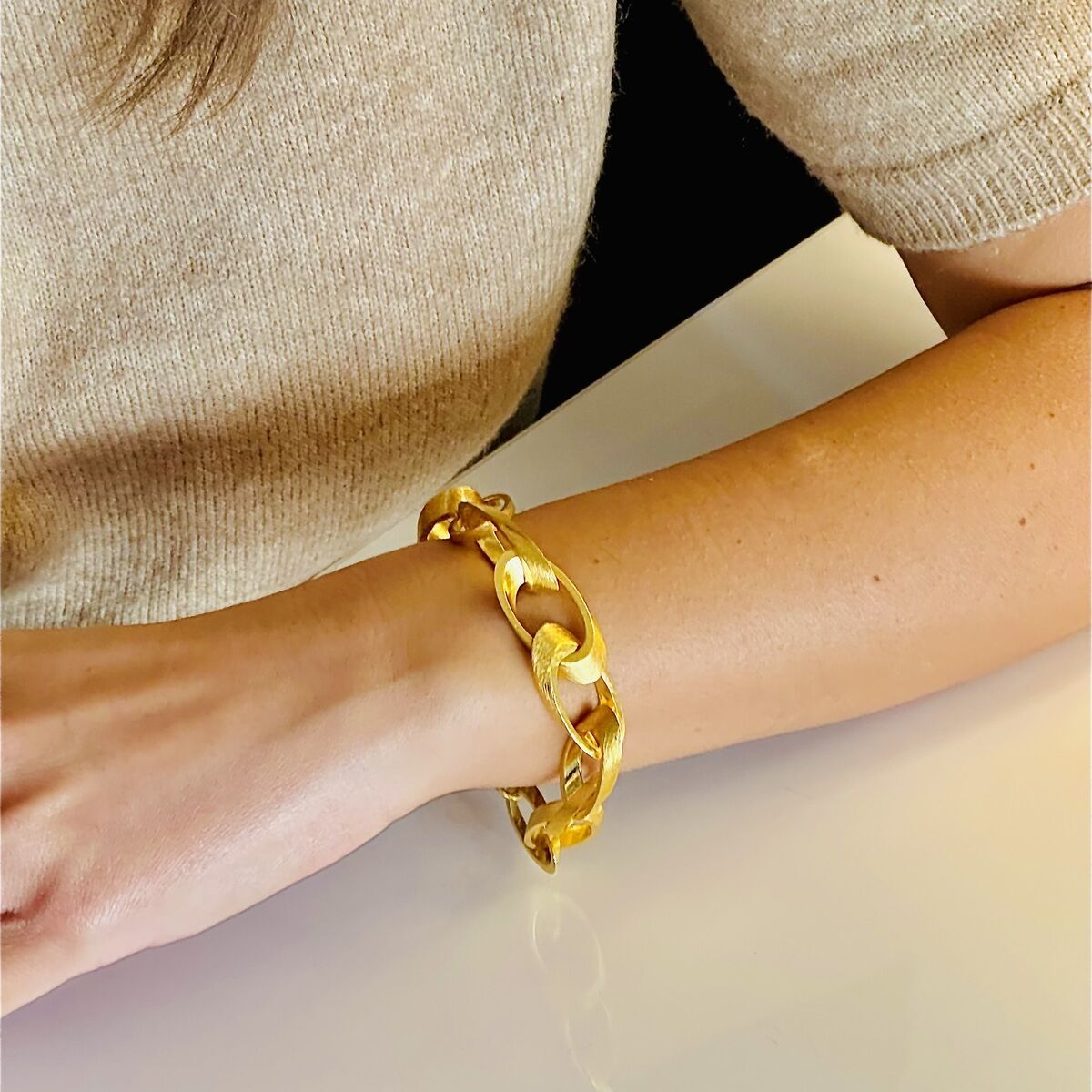 Nahaufnahme eines goldplattierten Silberarmbands am Handgelenk einer Person, die einen beigen Pullover trägt. Das Armband hat große Kettenglieder.