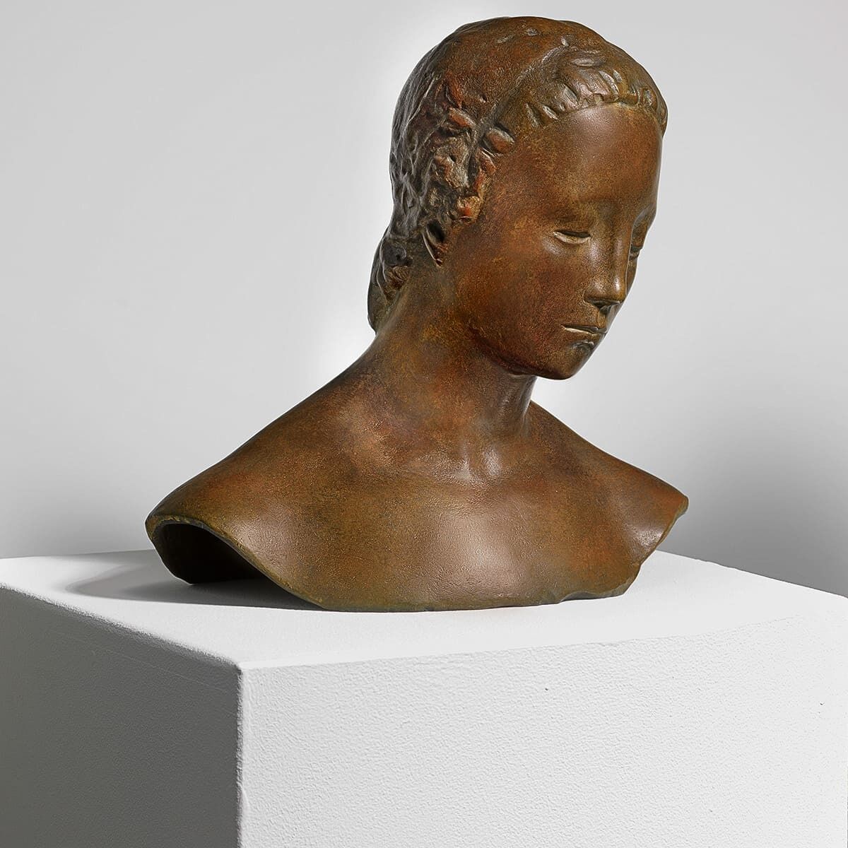 Büste einer Frau mit gesenktem Kopf, modelliert von Wilhelm Lehmbruck im Jahr 1910. Die Bronzeskulptur ist detailgetreu und zeigt geschlossene Augen und feine Gesichtszüge. Die Skulptur steht auf einem weißen Sockel vor einem neutralen Hintergrund.