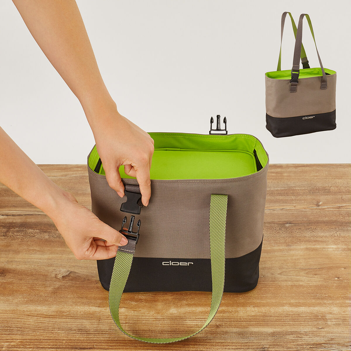 Eine Lunch-Tasche mit schwarzen, braunen und grünen Farbakzenten. Zwei Hände sind dabei, den Verschluss der Tasche zu öffnen. Die Tasche steht auf einem Holztisch.
