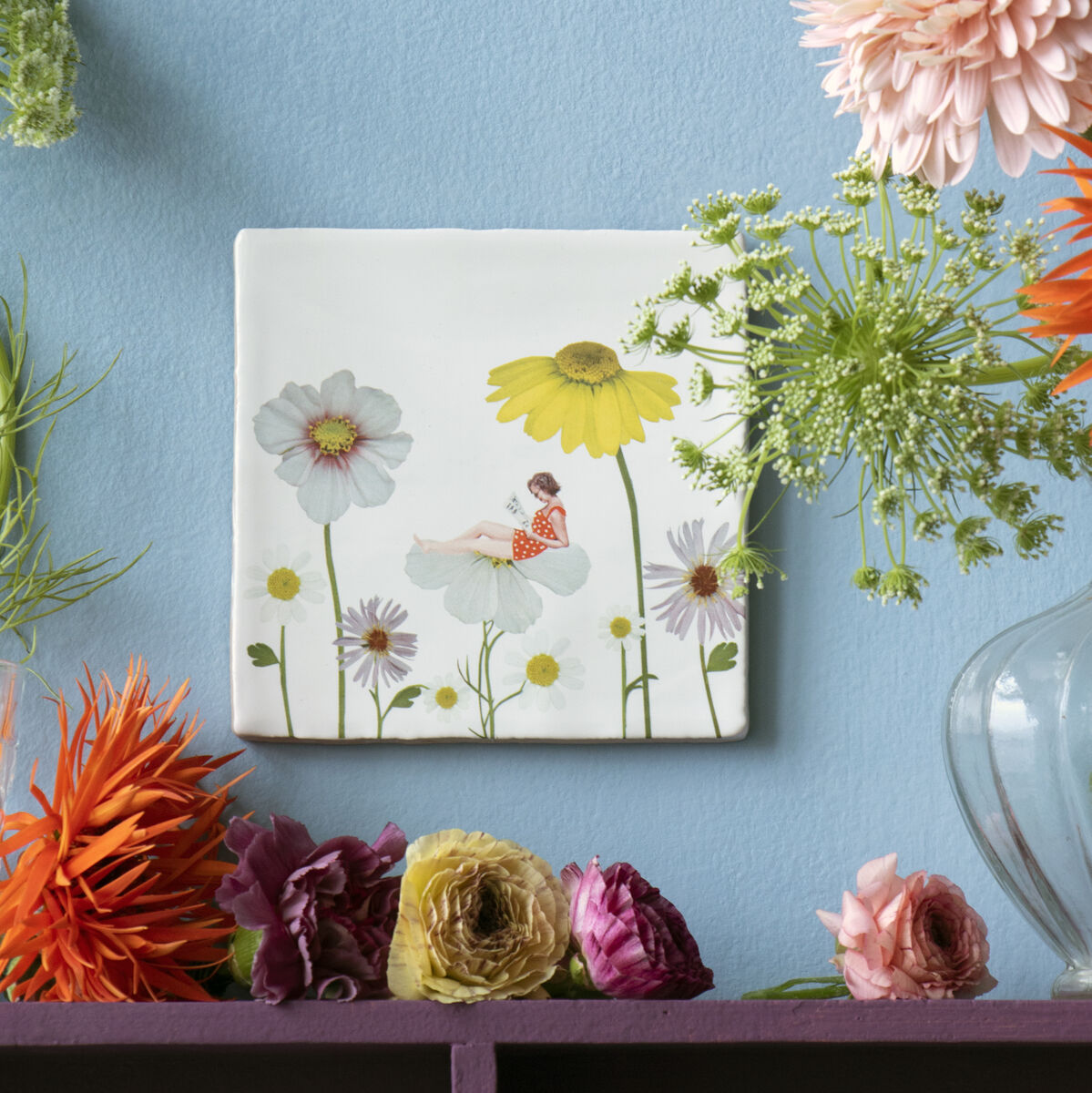 Keramikfliese mit floralem Design, platziert auf einem blauen Hintergrund neben einer Vase mit bunten Blumen wie orangefarbenen Dahlien, grünen und rosa Blüten. Perfekte Frühlings- oder Sommerdekoration für das Zuhause.