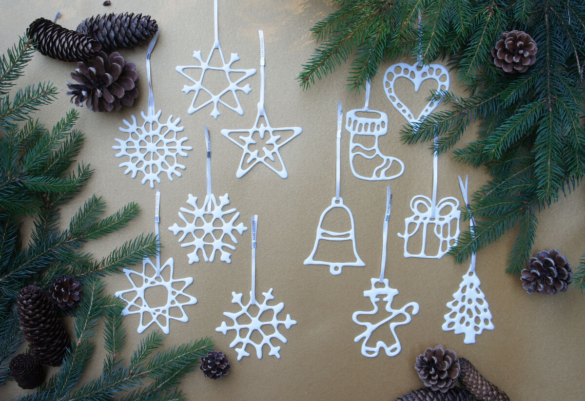 Weihnachtsanhänger-Set mit 6 weißen, unterschiedlichen Formen wie Schneeflocken, Baum, Herz, Stern, Glocke und Handschuh. Sie liegen auf einer braunen Oberfläche, umgeben von grünen Tannenzweigen und braunen Tannenzapfen.