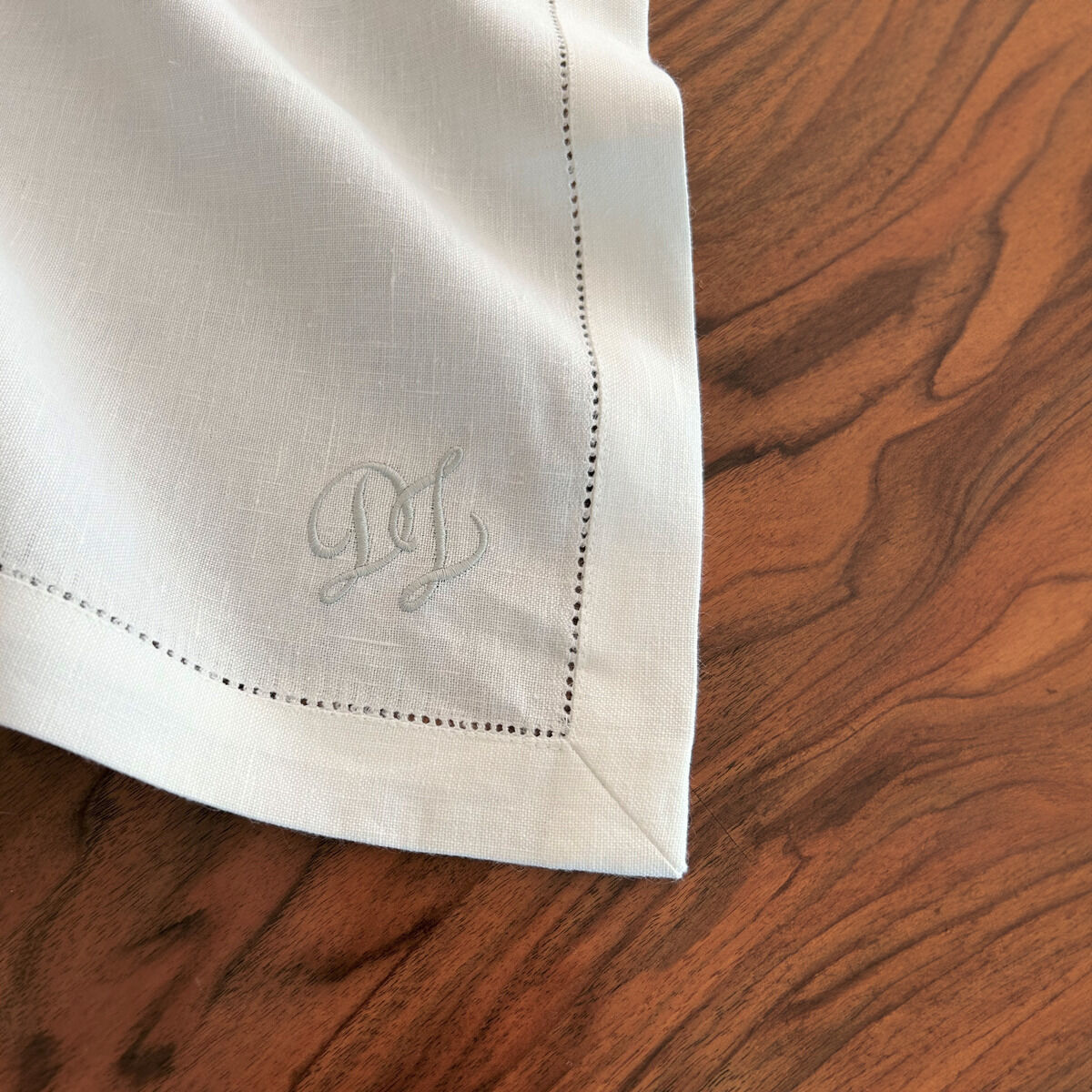 Leinenserviette in Weiß mit hellblauem Monogramm auf einem Holztisch. Die Serviette hat eine feine Stickerei an den Rändern.