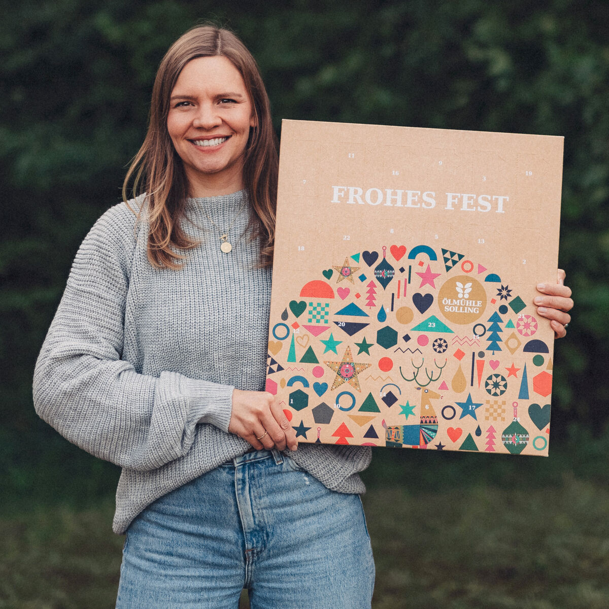 Eine lächelnde Frau steht im Freien vor einem grünen, bewaldeten Hintergrund und hält einen großen Adventskalender mit der Aufschrift 'Frohes Fest' sowie bunten weihnachtlichen Symbolen, wie Sterne, Tannen und Geschenke, in beiden Händen.
