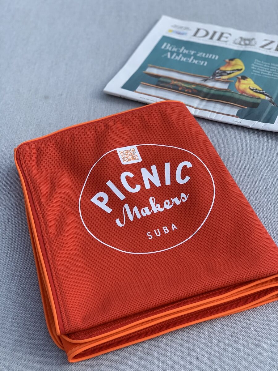 Eine orange Picknickdecke mit der Aufschrift 'PICNIC Makers SUBA' liegt zusammengefaltet auf einer grauen Oberfläche, neben einer Zeitung mit einem gelben Vogel auf dem Cover.