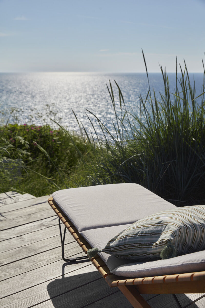 Ein komfortabler Liegestuhl mit weißer Auflage »MOLO« auf einer Holzterrasse mit Blick auf das Meer. Das Sonnenlicht glitzert auf dem Wasser, und im Hintergrund sind Strandhafer und grüne Pflanzen zu sehen. Perfekt für Entspannung und Außenbereich Dekoration.