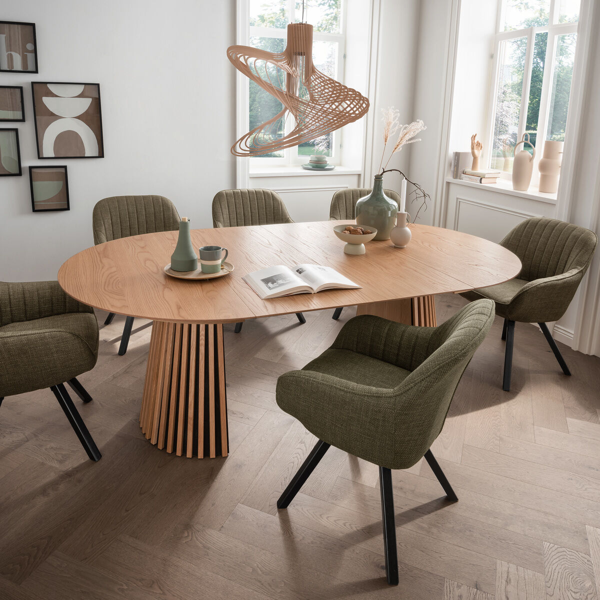 Ovaler Dining-Tisch "Oria 120cm" der Marke bene living steht im hellen Esszimmer. Der Tisch mit Naturholzoberfläche und gestreiftem Sockel ist von sechs olivgrünen, gepolsterten Stühlen umgeben. Auf dem Tisch liegen ein Buch, Tassen, Dekovasen und Keramikgeschirr.