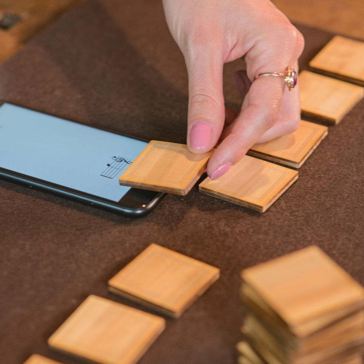 Hand legt ein Holzplättchen auf eine Oberfläche neben einem Smartphone mit Musiknoten. Mehrere Holzplättchen liegen verstreut auf einem Filzuntergrund.