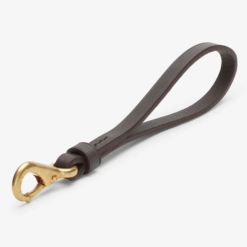 Auf hellem Hintergrund liegt ein eleganter Schlüsselanhänger von MISMO aus dunkelbraunem, pflanzlich gegerbtem Rindsleder mit sauber polierten Rändern. Der massive Karabinerhaken ist in matter goldener Optik gehalten und hebt sich kontrastreich vom dunklen Leder ab.