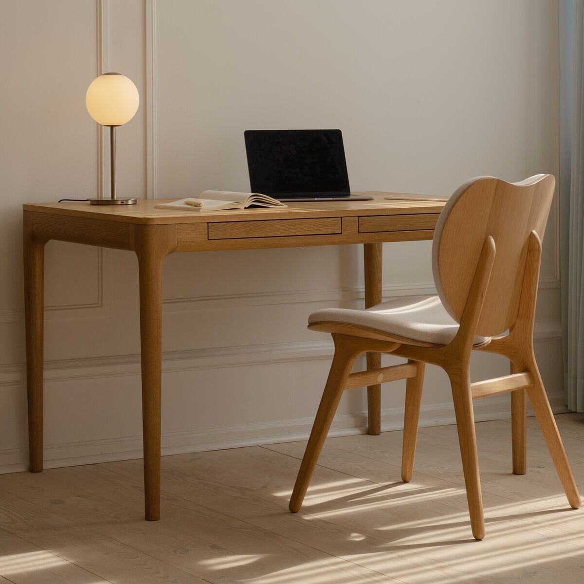 Heller, eleganter Arbeitsbereich mit Schreibtisch »Heart'n'Soul« von Umage aus naturfarbenem Eichenholz, darauf Laptop, Buch und Lampe mit runder, weißer Leuchte. Davor ein skandinavischer Stuhl mit heller Sitzfläche. Minimalistische, gemütliche Atmosphäre.