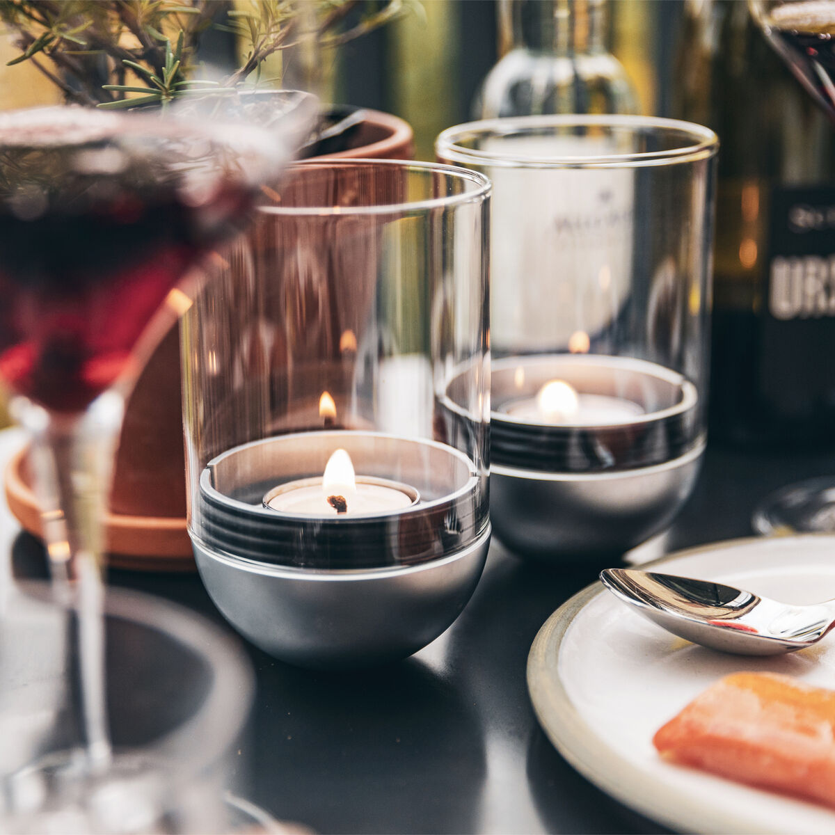 Ein eleganter Tisch mit verschiedenen Glaskerzenhaltern, die kleine brennende Teelichter halten. Daneben stehen halbvolle Weingläser und ein Teller mit Lachs. Die Szene vermittelt eine gemütliche und romantische Atmosphäre.