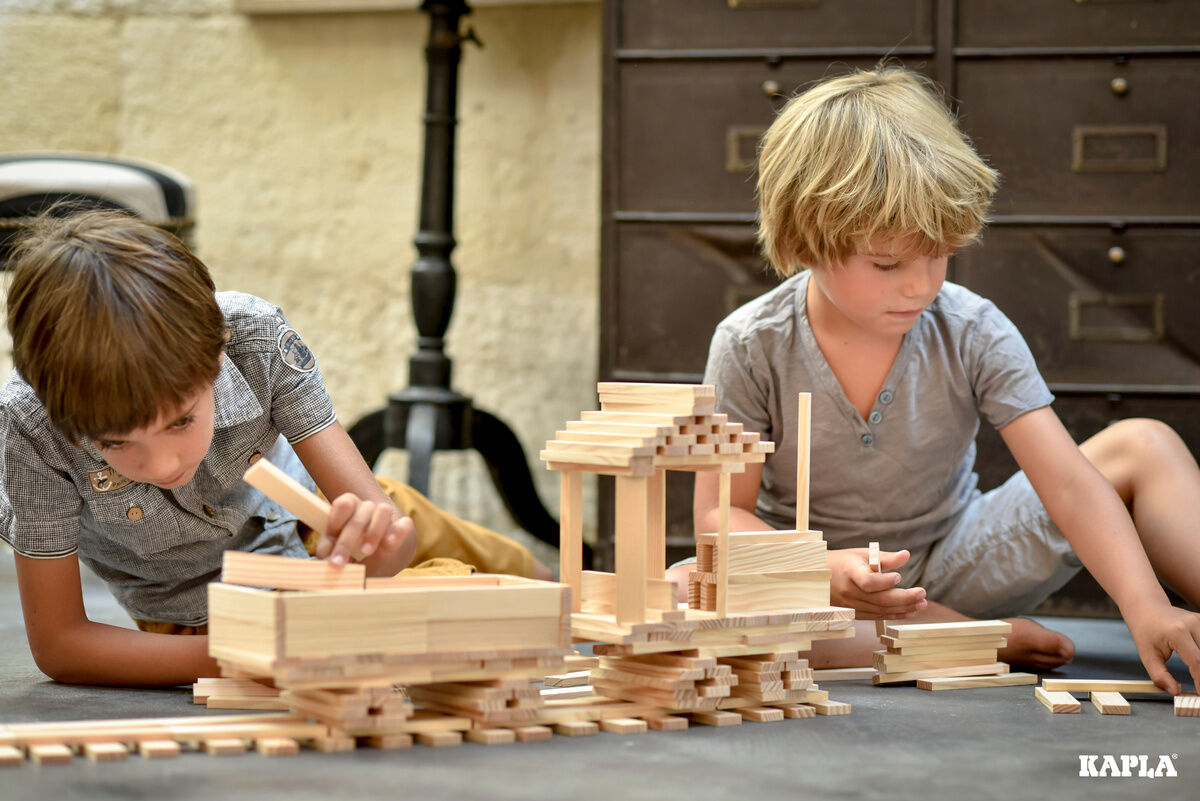 Zwei Kinder spielen mit Holzbausteinen und bauen eine Struktur auf einem dunklen Boden.