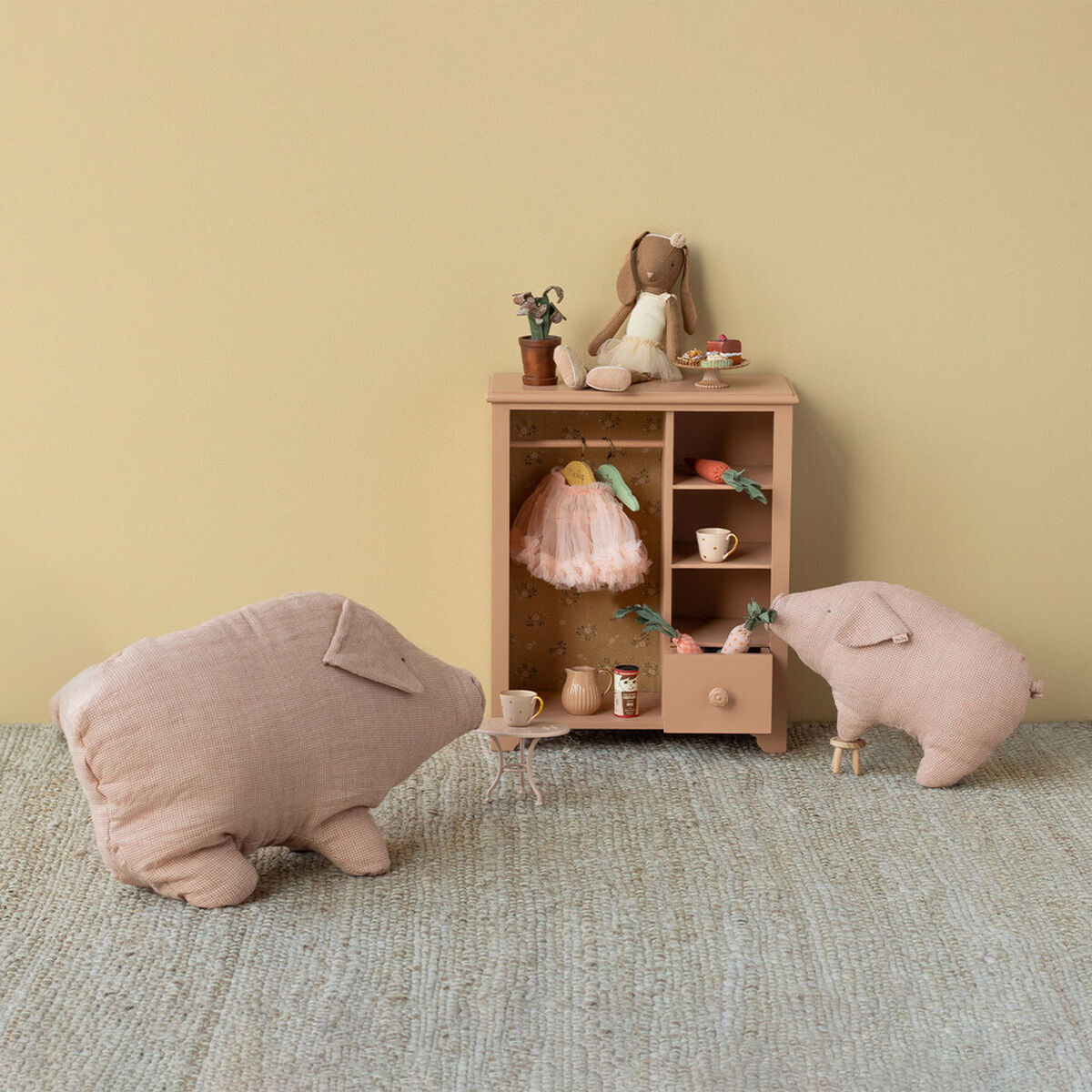 Das Bild zeigt zwei rosa Kuscheltiere „Schwein Polly Mittel“ der Marke Maileg aus Leinen/Baumwolle vor einem kleinen Puppenschrank mit Miniatur-Geschirr, einer Puppe und Dekoration. Der Hintergrund ist beige, der Boden grau-beige, das Gesamtambiente wirkt freundlich und einladend.