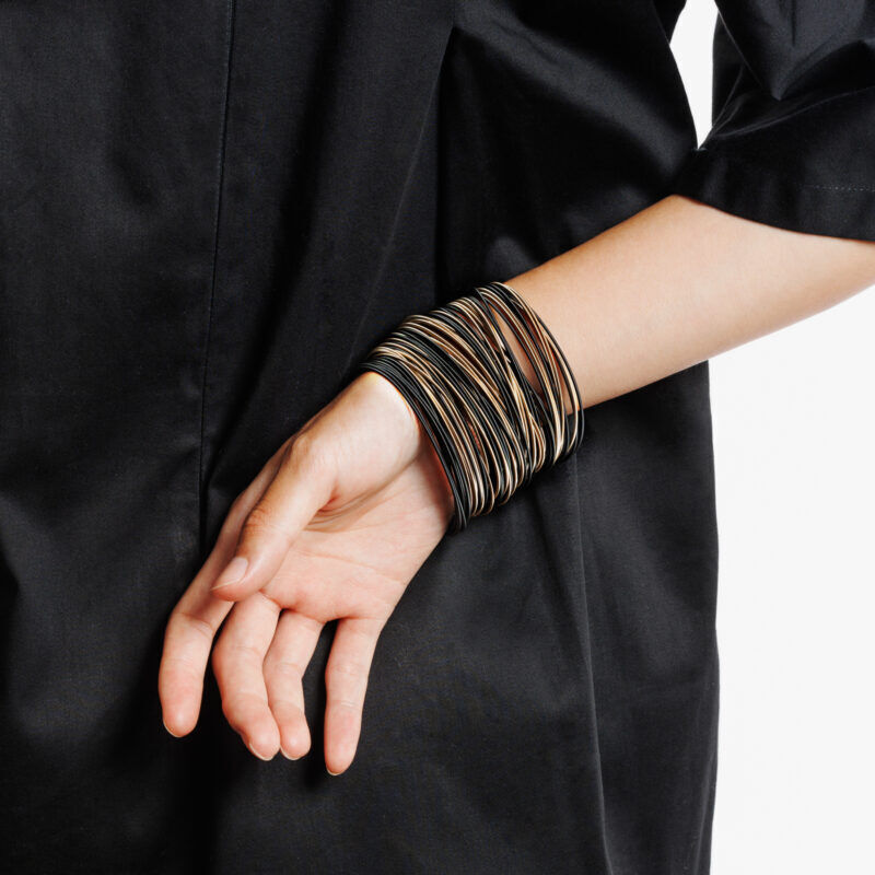 Ein schlankes Handgelenk trägt das Armband »Bronze & Black« von la molla, bestehend aus vielen einzelnen, elastischen Bändern in Bronze- und Schwarztönen. Das Armband liegt geschmeidig auf schwarzer Kleidung und setzt einen modernen Akzent.