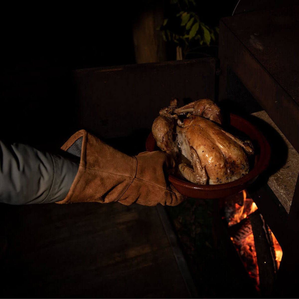 Ein Paar Handschuhe aus Rindleder, das beim Herausnehmen eines gebratenen Huhns aus einem Holzofen getragen wird. Die Szene ist in eine dunkle Umgebung mit einem offenen Feuer im Hintergrund gesetzt.